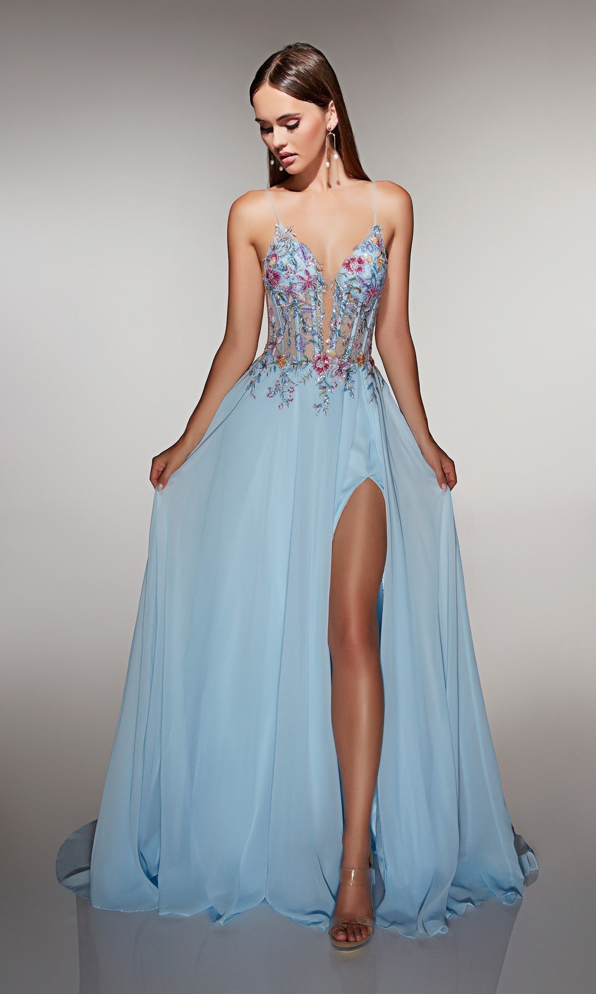 A woman in the Alyce 62179 long prom dress—light blue with a sheer, floral-embroidered corset bodice, thin straps, and a high slit—pairs it with open-toe heels and poses against a plain background.